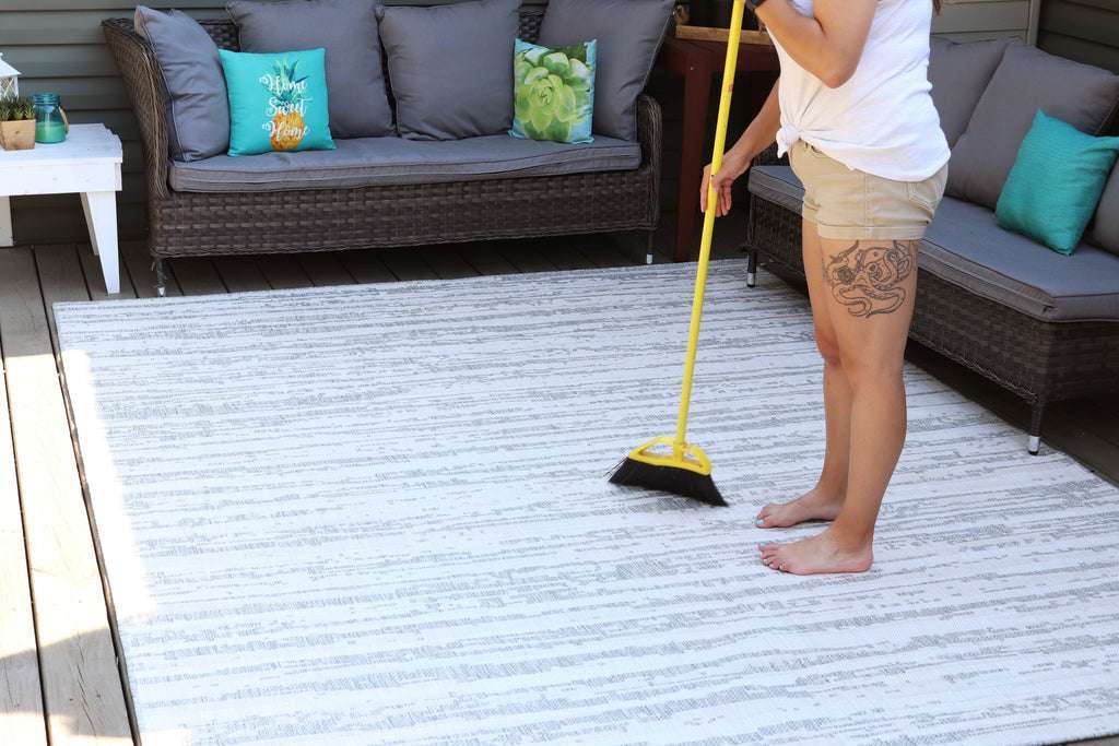 Person sweeping a striped grey and white outdoor rug on a wooden deck.